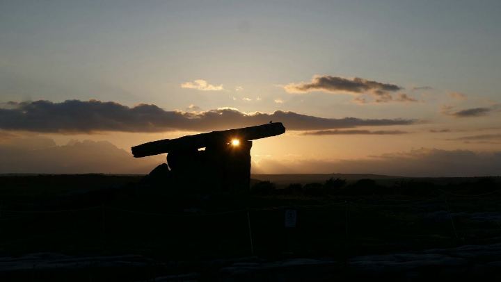 Un dolmen en silhouette contre un ciel de couchant ; le soleil passe sous la dalle.